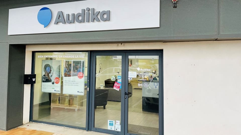 La façade d'une clinique Audika affiche le logo bleu et gris de l'entreprise. Texte visible : « BIEN ENTENDRE, ÇA CHANGE LA VIE ». L'intérieur montre des sièges et des affiches promouvant des appareils auditifs et services d'audiologie.