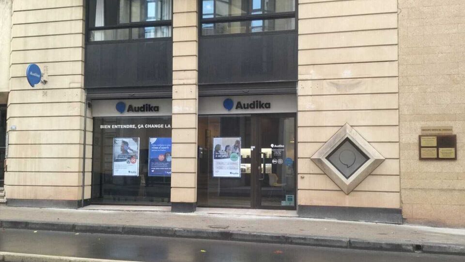 La façade d'une clinique auditive Audika avec deux vitrines affichant le slogan « Bien entendre, ça change la vie » et des posters promotionnels pour aides auditives et bilan auditif gratuit.