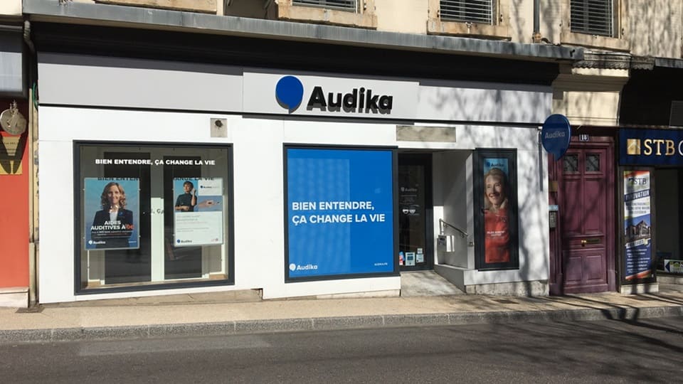 La façade d'une clinique Audika affiche 'BIEN ENTENDRE, ÇA CHANGE LA VIE' sur un panneau bleu. Des vitrines présentent des affiches publicitaires sur les aides auditives et l'accompagnement audiologique.