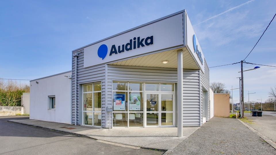 Le bâtiment principal est une clinique Audika spécialisée en soins auditifs. L’enseigne ’Audika’ est bien visible sur la façade en métal. Environnement urbain avec un ciel dégagé.