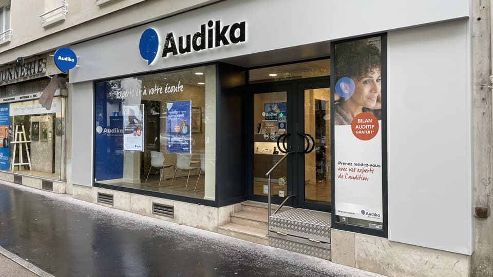 La façade d'une clinique Audika avec enseigne et logo bleus, affiches promotionnelles (ex. « BIEN ENTENDRE, ÇA CHANGE LA VIE »). Texte visible : « BILAN AUDITIF GRATUIT ».