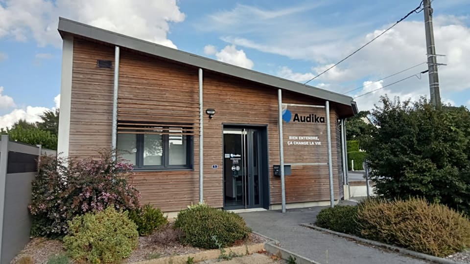 Bâtiment en bois avec logo ’Audika’ et slogan ’Bien entendre, ça change la vie’ sur la façade. Entrée vitrée, entourée de buissons et ciel bleu parsemé de nuages.
