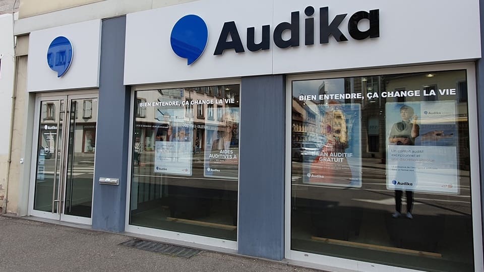 La façade d'une clinique Audika affiche le logo bleu et noir, des portes vitrées, et des posters annonçant : « Bien entendre, ça change la vie », « Bilan auditif gratuit ».