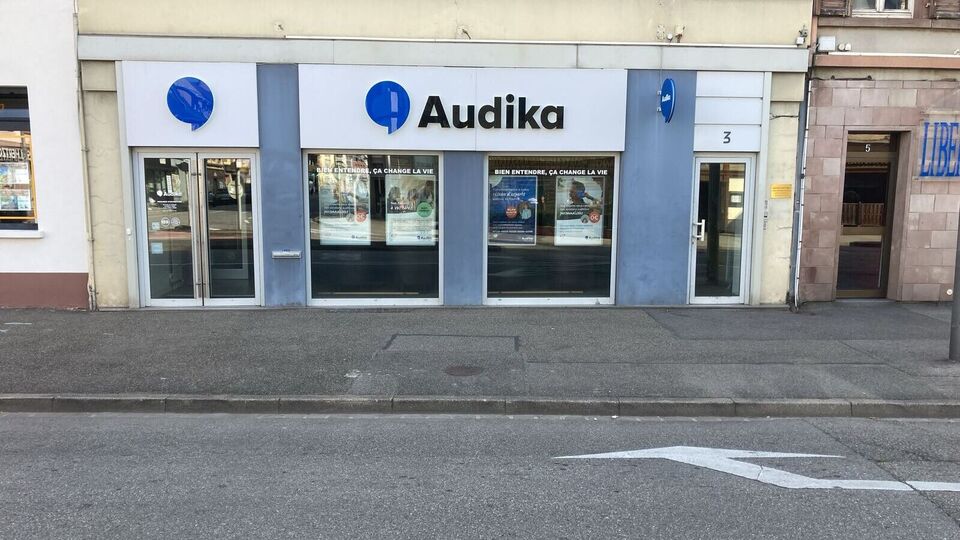 La façade d’une clinique Audika affiche le logo bleu et noir, des portes vitrées, et des posters annonçant : « Bien entendre, ça change la vie », « Bilan auditif gratuit ».