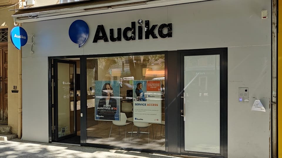 La devanture d'une clinique Audika affiche le logo bleu et noir de l’entreprise, avec des affiches promotionnelles sur les aides auditives et services d'audiologie visibles à travers les vitrines.