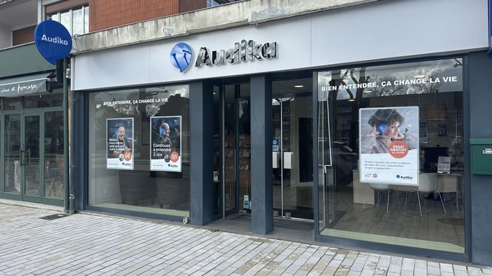 La façade d'une clinique Audika spécialisée en soins auditifs affiche le slogan « Bien entendre, ça change la vie » et des publicités mettant en avant des essais gratuits d'aides auditives. Le logo Audika est visible, et les vitrines présentent des images de patients avec des solutions auditives. Le contexte est urbain, avec un trottoir pavé devant l'entrée et des bâtiments adjacents.