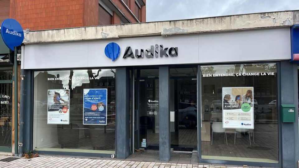 La façade d’une clinique Audika spécialisée en soins auditifs affiche le slogan « Bien entendre, ça change la vie » et des publicités mettant en avant des essais gratuits d’aides auditives. Le logo Audika est visible, et les vitrines présentent des images de patients avec des solutions auditives. Le contexte est urbain, avec un trottoir pavé devant l’entrée et des bâtiments adjacents.