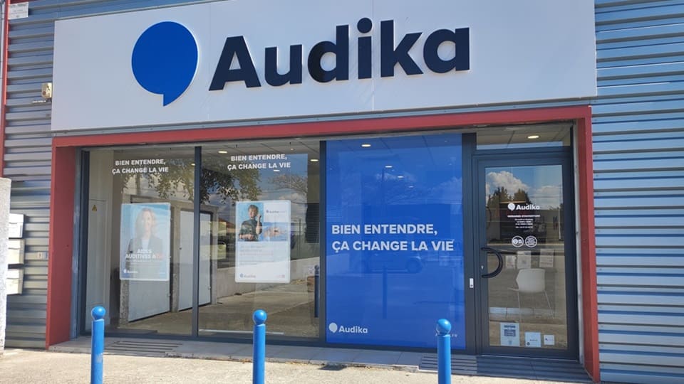 La façade d'une clinique Audika affiche le logo de la marque et le slogan « Bien entendre, ça change la vie » sur une vitrine vitrée, entourée d'un environnement urbain.