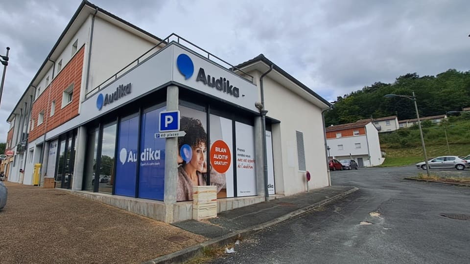 Un centre Audika est situé au coin d’un bâtiment moderne, affichant le texte : « Bilan auditif gratuit ». Contexte : rue calme avec des maisons, arbres et voitures.