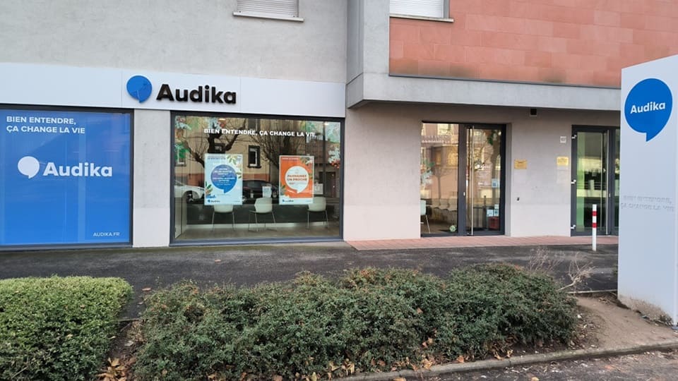 La façade d’une clinique Audika affiche le logo bleu et blanc avec le texte « Bien entendre, ça change la vie ». Les vitrines présentent des affiches colorées et l’intérieur visible montre une salle d’attente moderne. Un panneau Audika est situé dans un espace extérieur bordé de végétation.