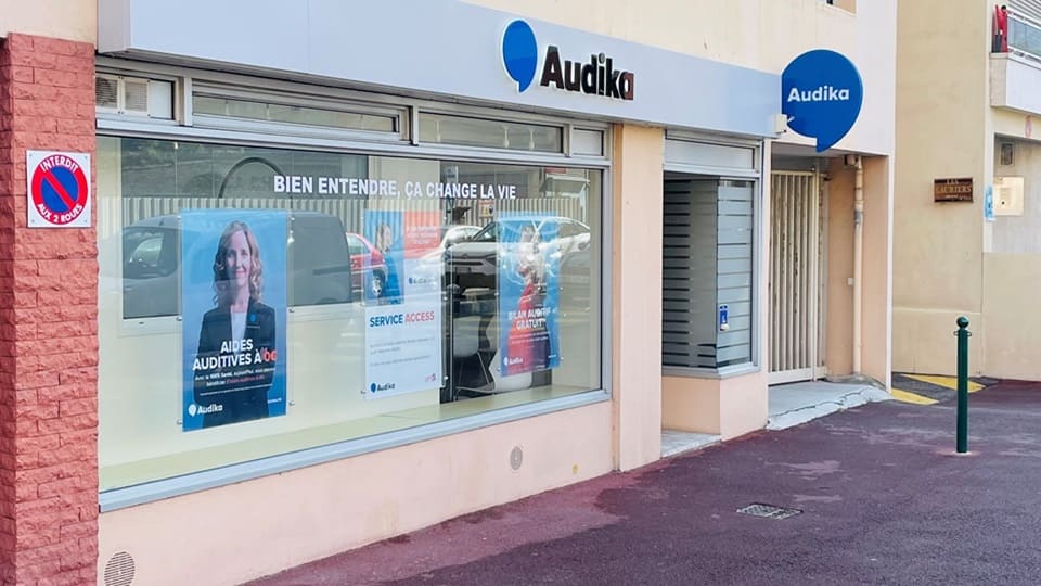La façade d'une clinique Audika affiche le slogan « Bien entendre, ça change la vie ». Des posters annoncent des « Aides auditives à 0€' », un « Bilan auditif gratuit », et le « Service Access ». Une signalisation « Interdit de stationner » est visible à gauche, et l'entrée de la clinique est située à droite, dans une rue urbaine.