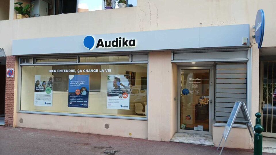 La façade d'une clinique Audika affiche le slogan « Bien entendre, ça change la vie ». Des posters annoncent des « Aides auditives à 0€' », un « Bilan auditif gratuit », et le « Service Access ». Une signalisation « Interdit de stationner » est visible à gauche, et l'entrée de la clinique est située à droite, dans une rue urbaine.