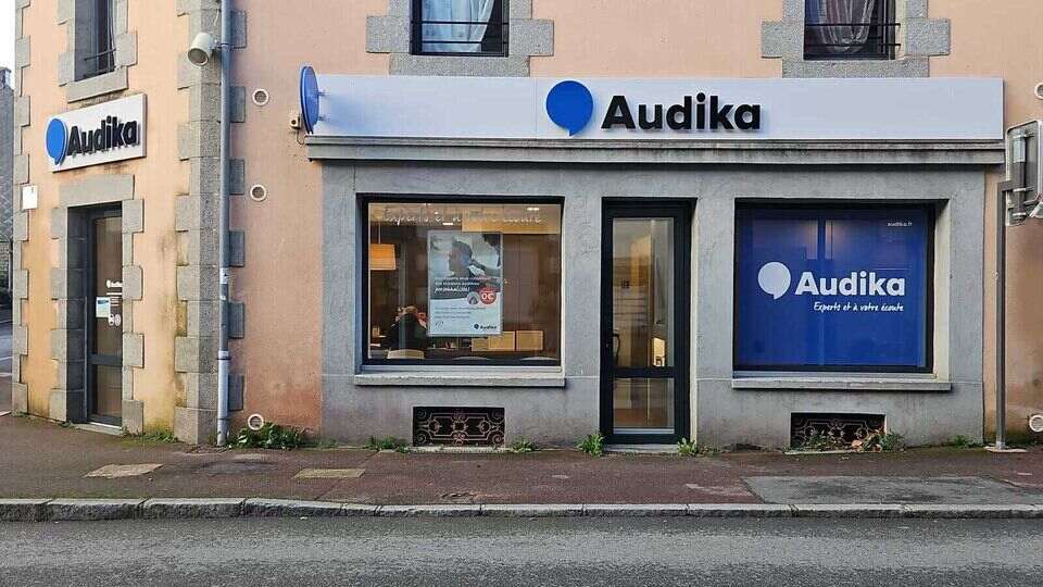 Façade d’une clinique Audika située dans un bâtiment en pierre beige avec encadrement en pierre grise. Une enseigne blanche affiche le logo 'Audika'. Texte visible : 'Bien entendre, ça change la vie'. Fleurissement suspendu sur un lampadaire rouge en premier plan.