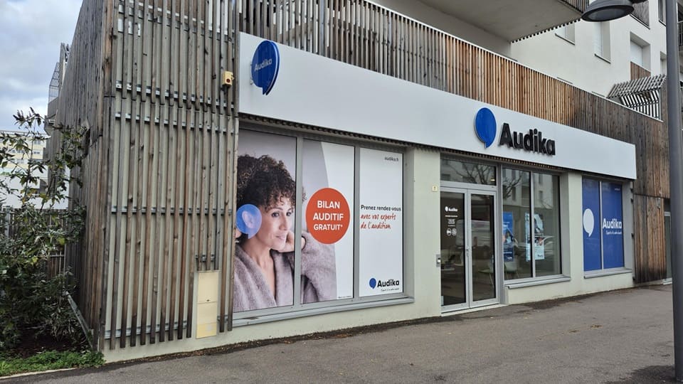 La façade d’une clinique Audika affiche le logo bleu caractéristique, avec les slogans : « Experts et à votre écoute » et « Bilan auditif offert’ ». Adresse web : audika.fr.