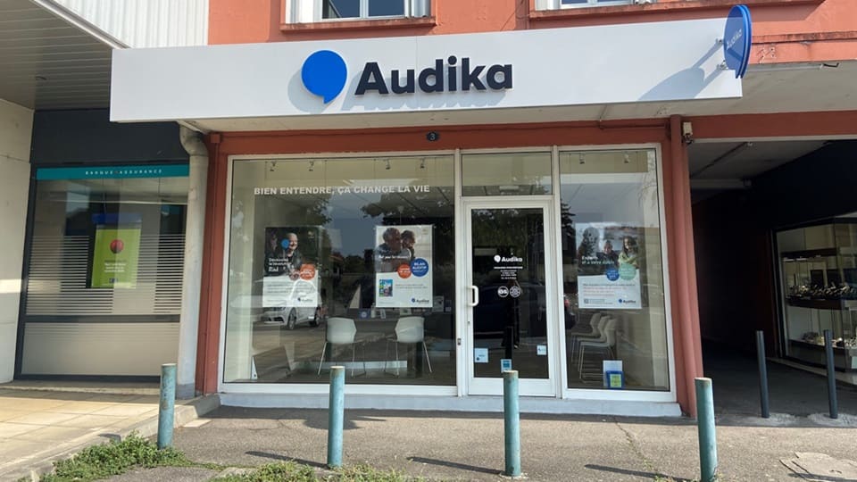 La façade d'une clinique Audika avec enseigne blanche et bleue, slogan « Bien entendre, ça change la vie », affiches promotionnelles, baies vitrées, et mobilier visible à l'intérieur.