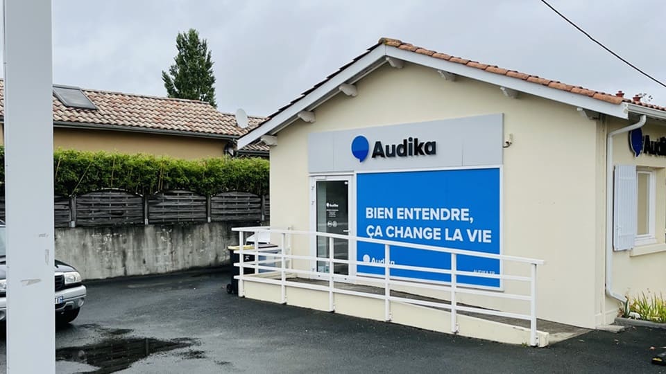 Un bâtiment beige avec le logo 'Audika' visible, affichant un panneau bleu : 'BIEN ENTENDRE, ÇA CHANGE LA VIE.' Lieu : parking avec barrière blanche.