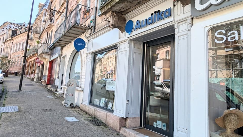 La façade d’un centre auditif Audika présente une vitrine et une porte en verre. Le contexte est une rue commerçante bordée de bâtiments anciens et de vitrines variées.