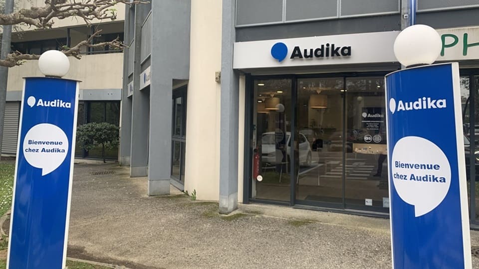 Deux colonnes bleues signalétiques avec le texte « Bienvenue chez Audika » encadrent l’entrée d’une clinique Audika, spécialisée en soins auditifs, située dans un immeuble moderne avec des baies vitrées.
