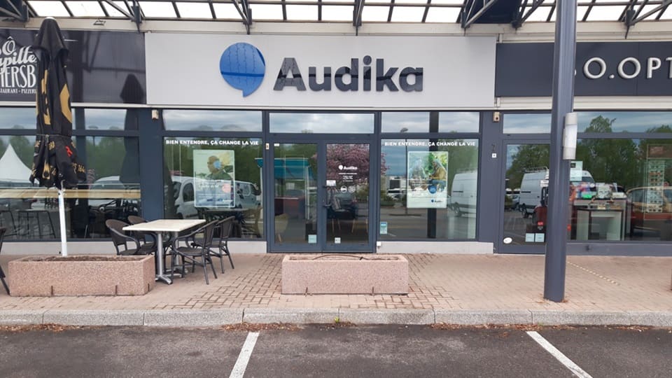 La façade d’un centre Audika, spécialisée dans les soins auditifs, affiche le slogan ’Bien entendre, ça change la vie’. Deux vitrines montrent des affiches, dans un environnement de zone commerciale.