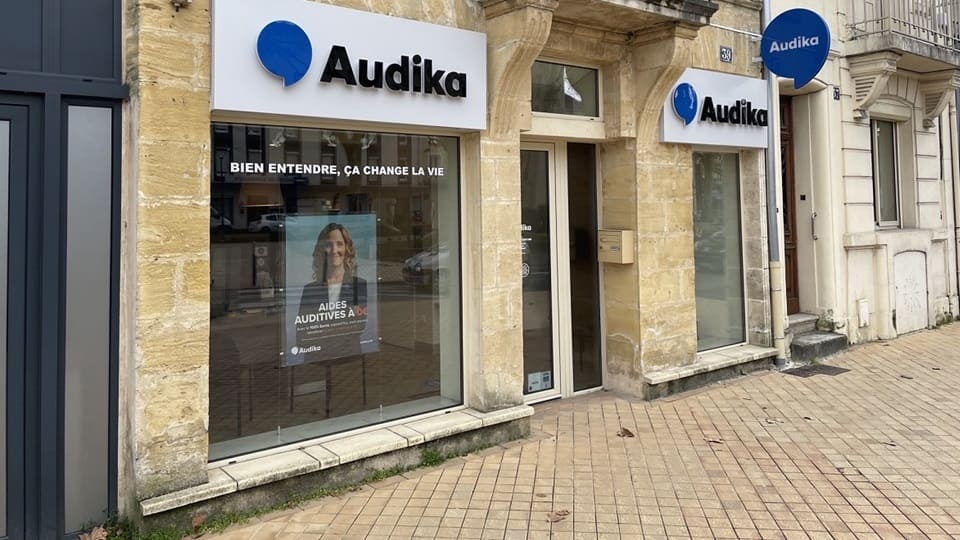 Façade d'une clinique Audika avec un panneau portant le logo bleu et le texte 'Audika'. Une vitrine affiche : 'BIEN ENTENDRE, ÇA CHANGE LA VIE' et une affiche mentionnant 'AIDES AUDITIVES'. Le bâtiment en pierre est situé dans une rue pavée.