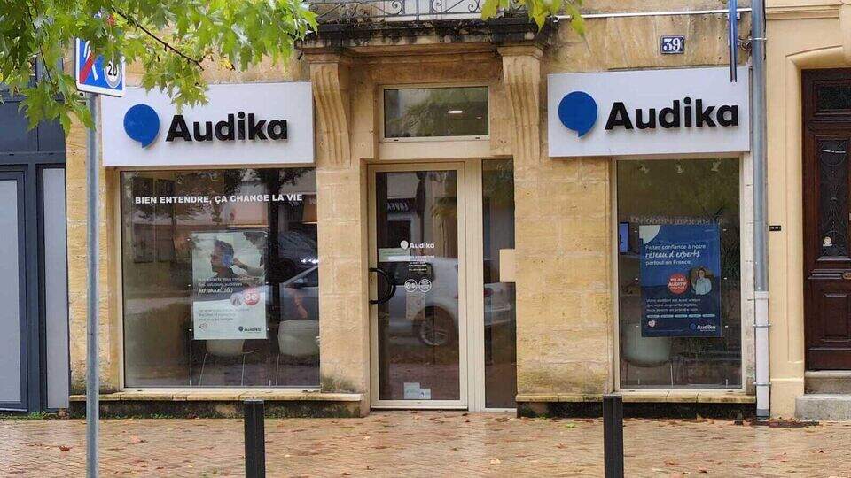 Façade d’une clinique Audika avec un panneau portant le logo bleu et le texte ’Audika’. Une vitrine affiche : ’BIEN ENTENDRE, ÇA CHANGE LA VIE’ et une affiche mentionnant ’AIDES AUDITIVES’. Le bâtiment en pierre est situé dans une rue pavée.