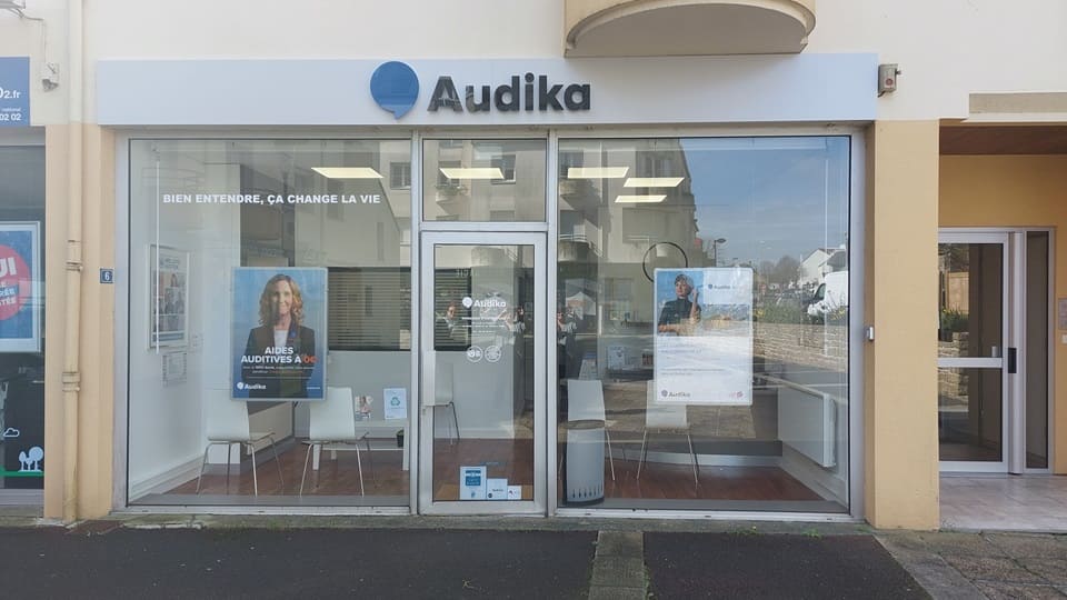 La façade d'un centre Audika spécialisé en soins auditifs arbore des vitrines transparentes avec affiches publicitaires mentionnant « BIEN ENTENDRE, ÇA CHANGE LA VIE » et « AIDES AUDITIVES ». L'intérieur lumineux montre des chaises et un bureau, situé dans un environnement urbain.