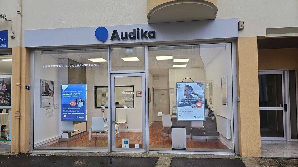 La façade d’un centre Audika spécialisé en soins auditifs arbore des vitrines transparentes avec affiches publicitaires mentionnant « BIEN ENTENDRE, ÇA CHANGE LA VIE » et « AIDES AUDITIVES ». L’intérieur lumineux montre des chaises et un bureau, situé dans un environnement urbain.