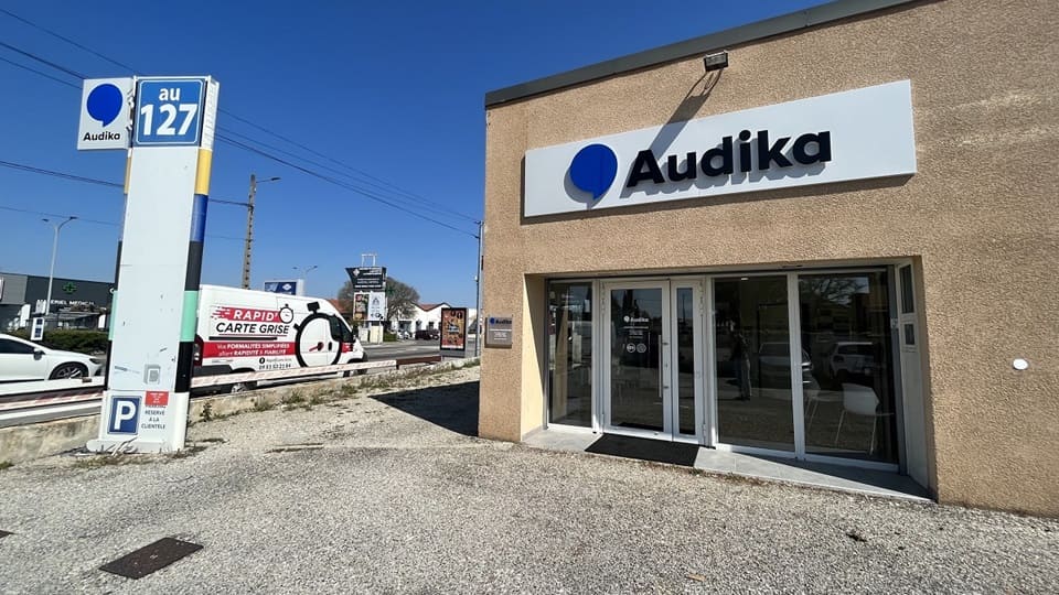 La façade d’un centre Audika, spécialisé en soins auditifs, affiche un grand logo et des portes vitrées. Contextuellement, un panneau indique ’au 127’ et un parking réservé est visible.
