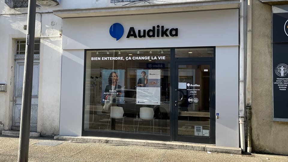 Une façade de clinique Audika avec vitrine où figurent le slogan « Bien entendre, ça change la vie » et des affiches publicitaires pour aides auditives, située en zone urbaine.