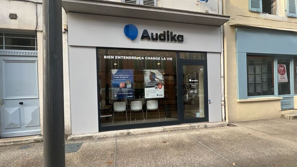 Une façade de clinique Audika avec vitrine où figurent le slogan « Bien entendre, ça change la vie » et des affiches publicitaires pour aides auditives, située en zone urbaine.