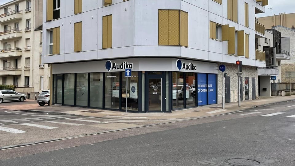 Une façade de clinique auditive Audika située à un carrefour urbain. Texte visible : 'BIEN ENTENDRE, ÇA CHANGE LA VIE.' Environnement : immeubles résidentiels modernes, passage piéton, trottoirs.