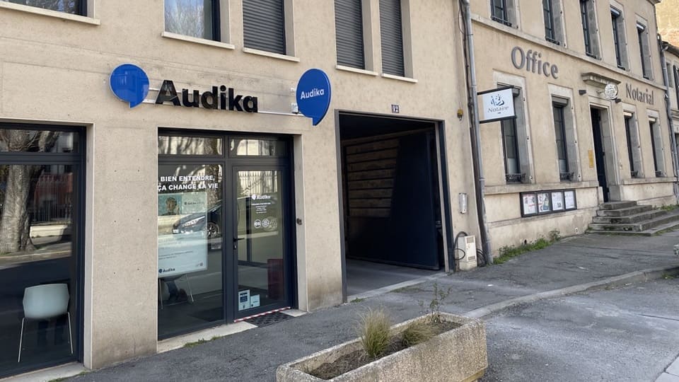La façade d'un centre auditif Audika affiche le logo bleu et noir, avec une vitrine portant le slogan 'Bien entendre, ça change la vie'. À droite, un bâtiment indique 'Office Notarial'.