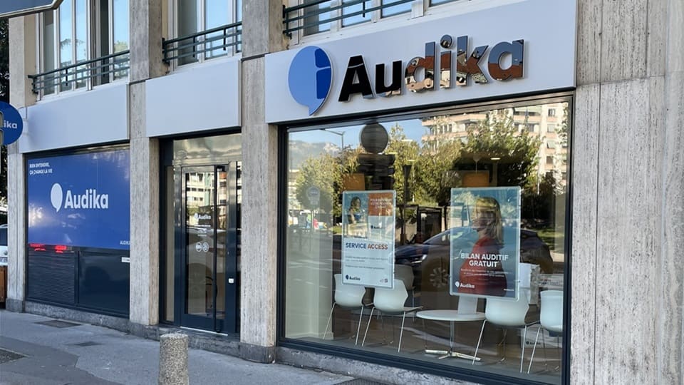 La façade d’une clinique Audika présente un logo identifiable avec ses vitrines affichant des posters : « SERVICE ACCESS » et « BILAN AUDITIF GRATUIT ». Environnement urbain en arrière-plan.