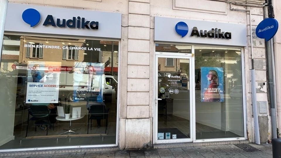 La façade d'une clinique Audika arbore le logo bleu et noir de l'entreprise, avec les slogans 'BIEN ENTENDRE, ÇA CHANGE LA VIE' et 'BILAN AUDITIF GRATUIT' affichés sur les vitrines. À l’intérieur, des affiches vantent les 'AIDES AUDITIVES' et les 'SERVICE ACCESS,' reflétant les services dédiés à l'audiologie et aux appareils auditifs.