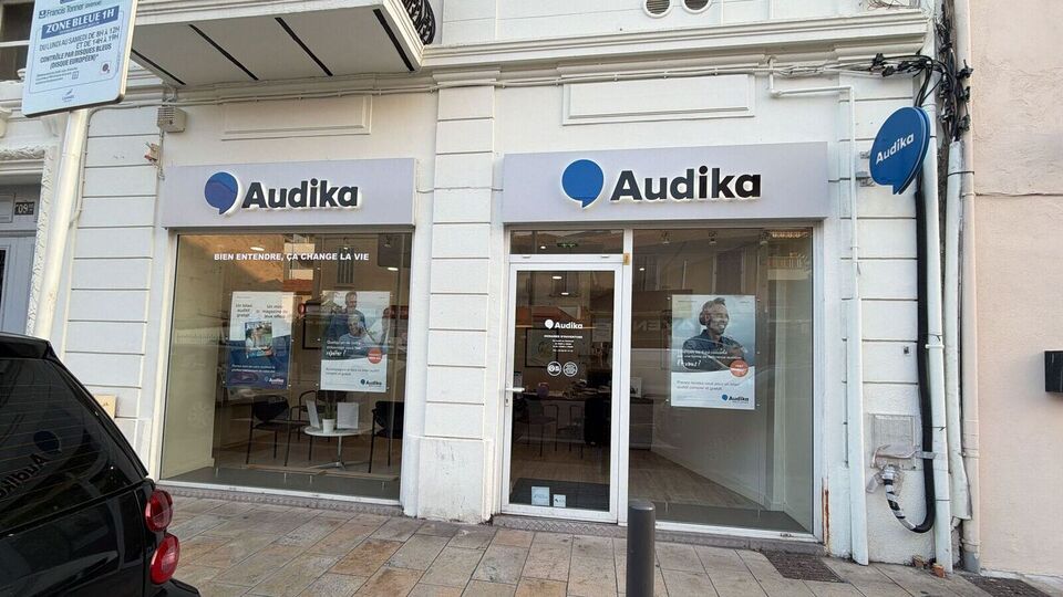 La façade d’une clinique Audika arbore le logo bleu et noir de l’entreprise, avec les slogans ’BIEN ENTENDRE, ÇA CHANGE LA VIE’ et ’BILAN AUDITIF GRATUIT’ affichés sur les vitrines. À l’intérieur, des affiches vantent les ’AIDES AUDITIVES’ et les ’SERVICE ACCESS,’ reflétant les services dédiés à l’audiologie et aux appareils auditifs.