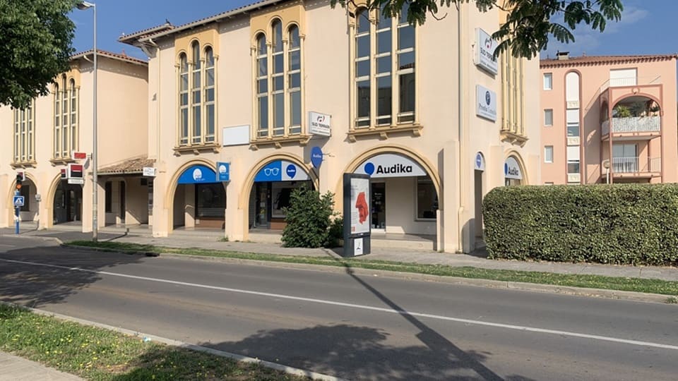 Façade d'un centre Audika, spécialisé dans les aides auditives et tests auditifs, situé dans un bâtiment aux arcs beige clair avec fenêtres verticales, jouxtant une rue bordée d'arbres et de buissons.
