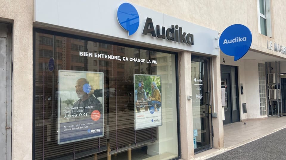 Une façade de clinique Audika avec vitrines affichant deux publicités :  
            1) Texte : « Découvrez nos produits quasi invisibles à partir de 0€'. N'attendez plus pour retrouver un vrai confort d'écoute. Audika ».  
            2) Texte : « Audika Blu Max. Nouvelle génération d’aides auditives rechargeables. Audika ».   
            Slogan vitre : « Bien entendre, ça change la vie ».  
            Environnement urbain calme.