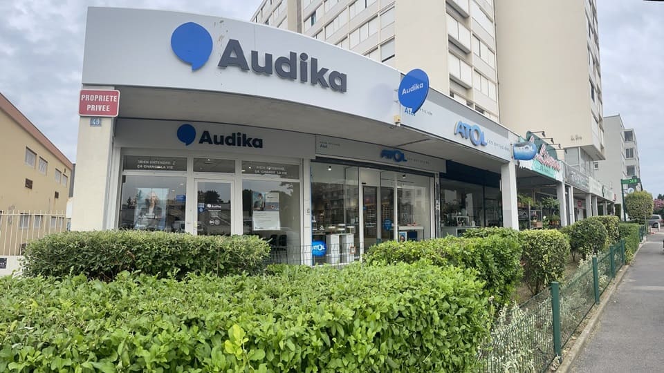 La façade de la clinique Audika, spécialisée en soins auditifs, affiche un logo bleu, entourée d’arbustes. Texte visible : « Audika », « Propriété privée », et « ATOL Les Opticiens ».
