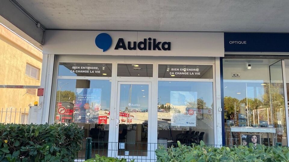 La façade de la clinique Audika, spécialisée en soins auditifs, affiche un logo bleu, entourée d’arbustes. Texte visible : « Audika », « Propriété privée », et « ATOL Les Opticiens ».