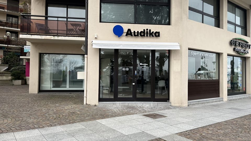 La façade d’une clinique Audika présente un logo bleu et noir, avec des portes vitrées et un store blanc. Le contexte inclut une place pavée et des bâtiments voisins.