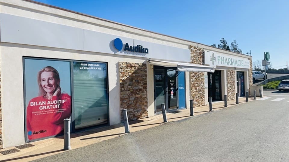 Une façade de centre auditif Audika avec une affiche indiquant 'BILAN AUDITIF GRATUIT' et 'BIEN ENTENDRE, ÇA CHANGE LA VIE,' à côté d'une pharmacie dans un environnement ensoleillé.