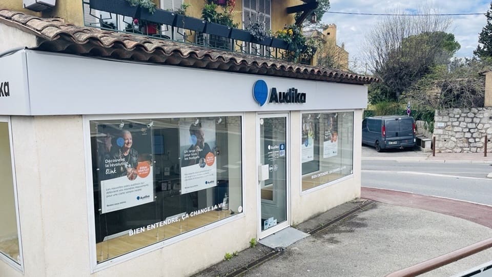La façade d’un centre auditif Audika, avec vitrines affichant des publicités sur les aides auditives et tests auditifs. Texte visible : ’Bien entendre, ça change la vie’ et ’Découvrez la révolution Blink.’ Contexte : rue calme avec végétation et immeubles résidentiels.