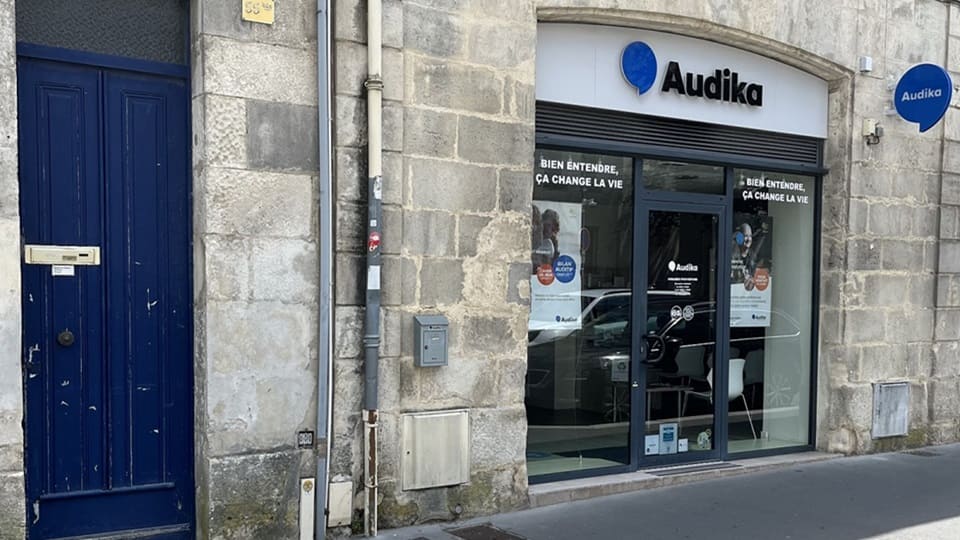 La façade d’un centre Audika, spécialisé en soins auditifs, affiche le logo bleu 'Audika' et le slogan « Bien entendre, ça change la vie ». Environnement urbain en pierre traditionnelle.