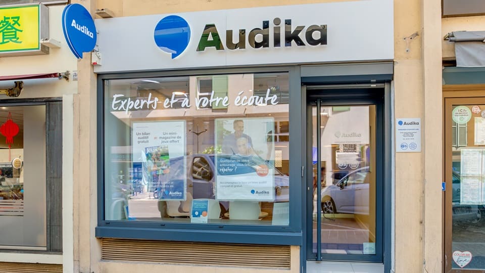 La façade d’un centre Audika spécialisé en santé auditive affiche un logo bleu et noir. À gauche, une vitrine lumineuse dévoile des espaces modernes; contextuellement, des commerces adjacents.