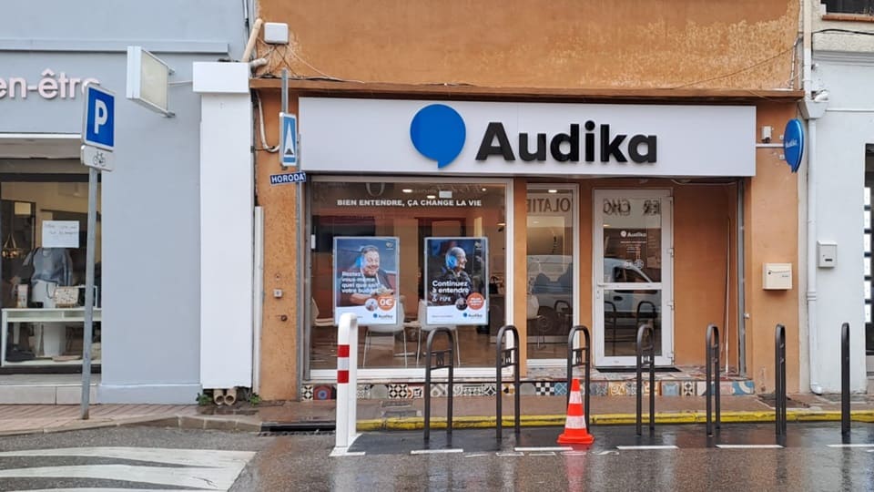 La façade d’un centre auditif Audika avec logo bleu, slogan ’Bien entendre, ça change la vie,’ affiches publicitaires et accès protégé par des poteaux, située dans une rue urbaine.
