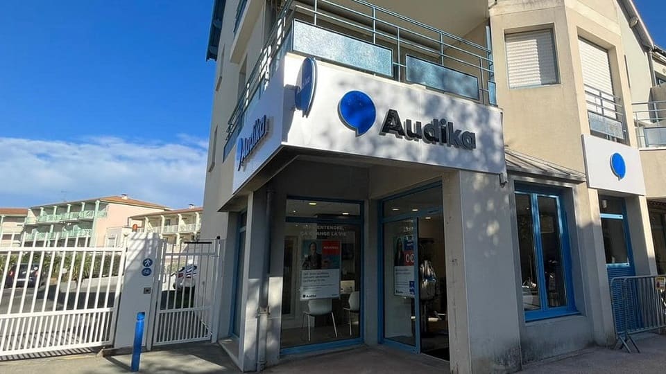 La façade d'une clinique Audika, spécialisée en soins auditifs, affiche un logo bleu. Des affiches à l'intérieur mentionnent des tests auditifs et offres. Environnement ensoleillé avec bâtiments résidentiels.