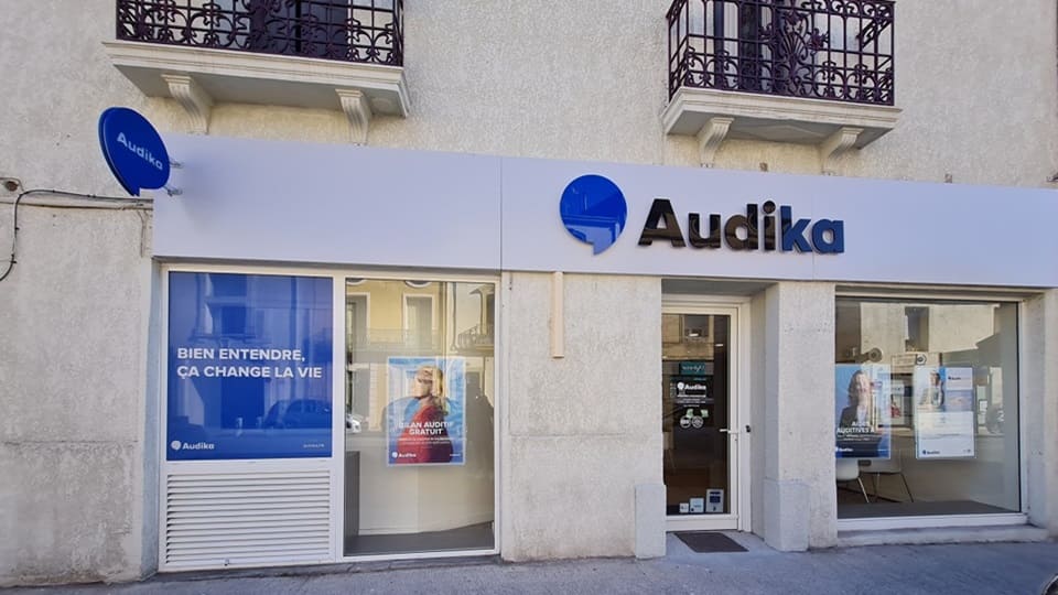La façade d'une clinique Audika affiche un logo bleu et noir, avec les textes 'BIEN ENTENDRE, ÇA CHANGE LA VIE' et 'UN AUDIT GRATUIT'. L'intérieur est visible à travers les vitrines.