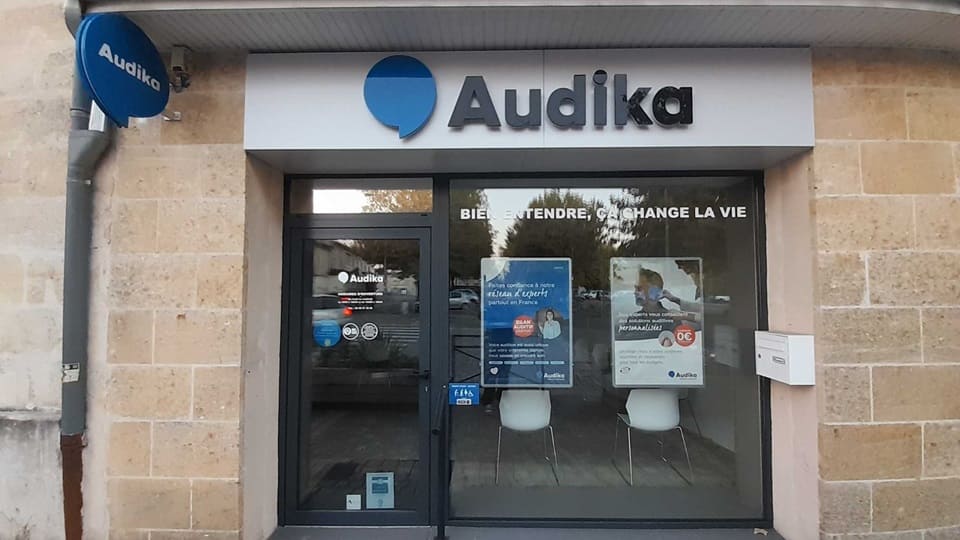 La façade d’un centre Audika avec une enseigne blanche et bleue, affichant le slogan ’Bien entendre, ça change la vie’. Deux affiches promotionnelles évoquent des offres sur les aides auditives avec ’100% Santé’ et ’Nouveauté’. À travers la vitrine, on distingue un intérieur lumineux avec des sièges blancs et une ambiance professionnelle. Les services incluent des tests auditifs et la vente d’appareils auditifs.
