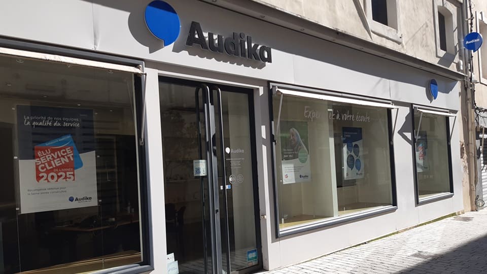 Façade d’un centre auditif Audika, avec vitrines montrant affiches sur les services, dont une mentionne : ’Élu Service Client de l’Année 2025.’ Environnement urbain pavé et lumineux.
