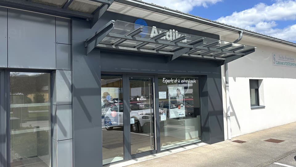La façade d’une clinique Audika présente des vitrines décorées de ballons colorés et d’affiches. Texte visible : « Experts et à votre écoute »